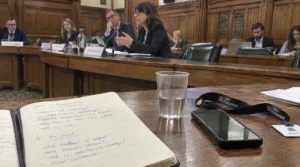 A candid photo from the audience of a parliamentary committee meeting in Portcullis House, Westminster, showing a notebook with handwritten questions on digital strategy next to a water glass and a UK Parliament visitor lanyard.