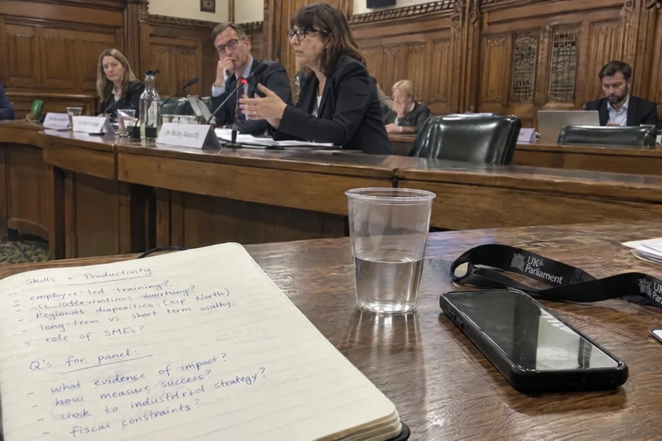 A candid photo from the audience of a parliamentary committee meeting in Portcullis House, Westminster, showing a notebook with handwritten questions on digital strategy next to a water glass and a UK Parliament visitor lanyard.